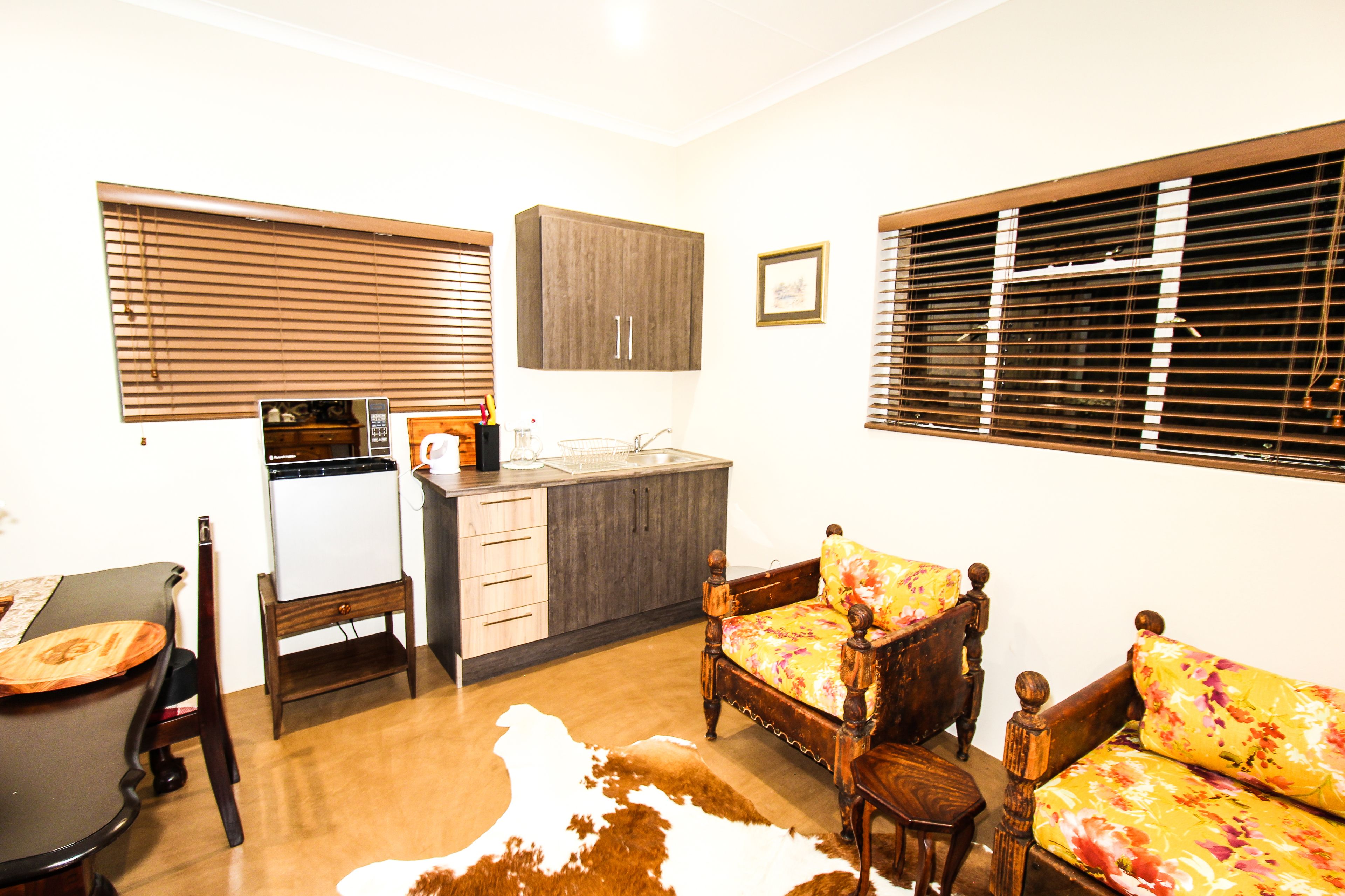 A compact kitchen with a microwave, sink, and wooden cabinets. There is a dining table with chairs and a sofa with floral upholstery.