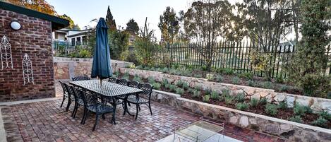 Un patio con suelo de ladrillo, una mesa y sillas de metal, una jardinera de piedra y una parrilla de metal.