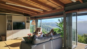 A modern living room with a large gray sofa, a flat-screen TV, and a view of a mountainous landscape through large glass doors. - Oubaai Villa  (Herolds Bay)