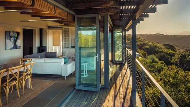 A modern outdoor living space with a wooden deck, a white sofa, and a dining area with wicker chairs. - Oubaai Villa  (Herolds Bay)