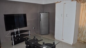A compact living space with a flat-screen TV, a refrigerator, a white wardrobe, and a glass coffee table. - Willu Executive Lodge (Livingstone)