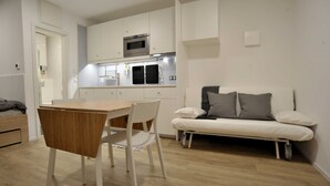 A compact kitchen with white cabinetry, a microwave, and a built-in oven. There is a dining table with chairs and a sofa with cushions. - Apartment 1 - FREE Ride cabins (Tösens)