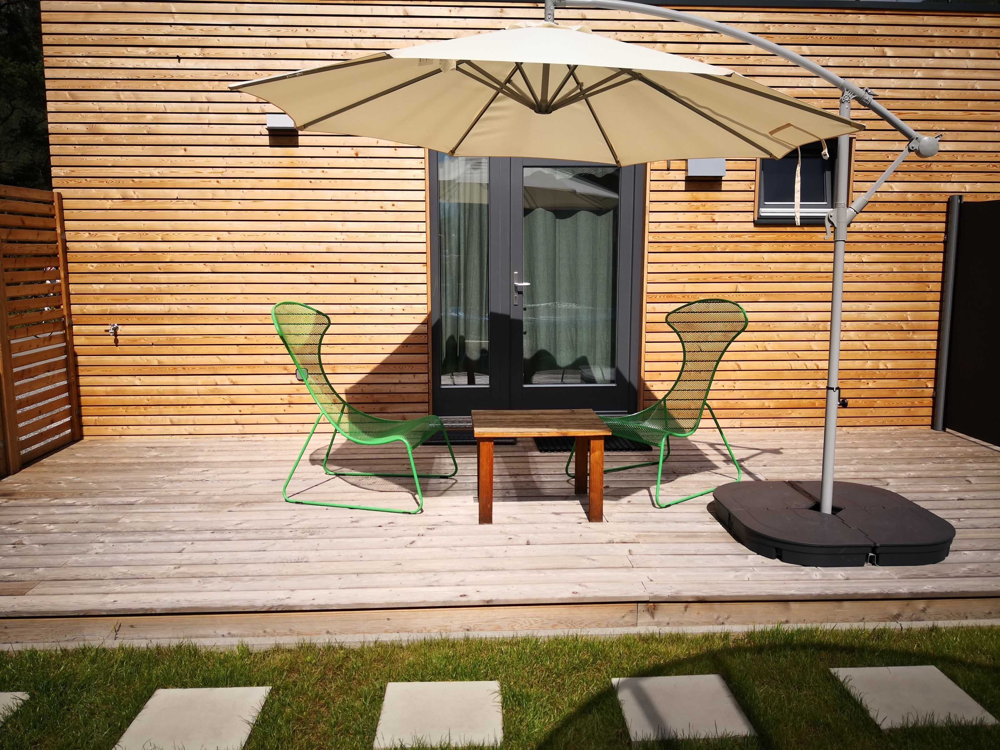 Eine Holzterrasse mit zwei grünen Stühlen und einem Tisch unter einem großen Sonnenschirm.