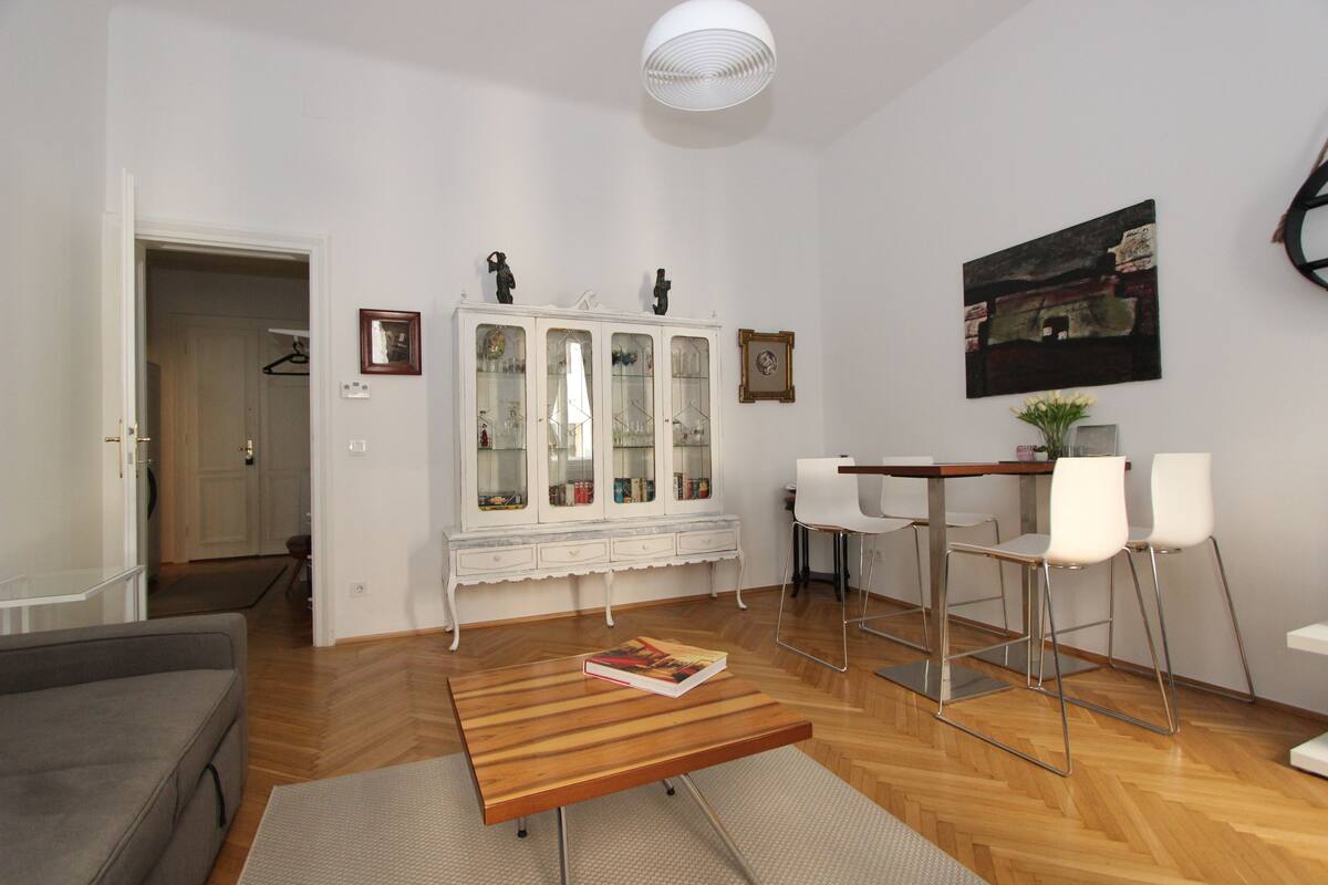 A living room with a wooden coffee table, a grey sofa, a white cabinet with glass doors, and a dining area with a white table and chairs.