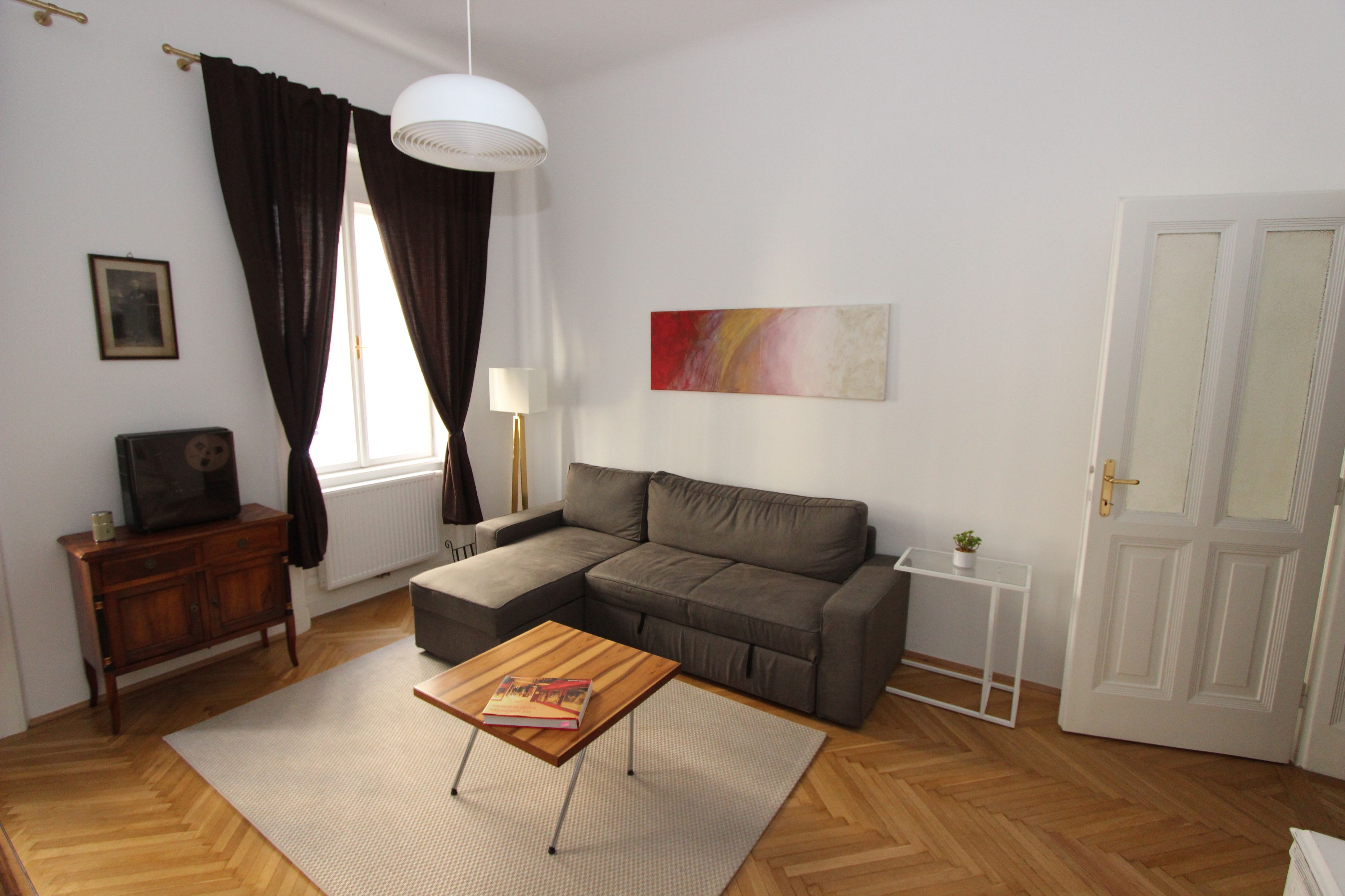 A living room with a sectional sofa, a wooden coffee table, a white lamp, a framed picture, and a door.