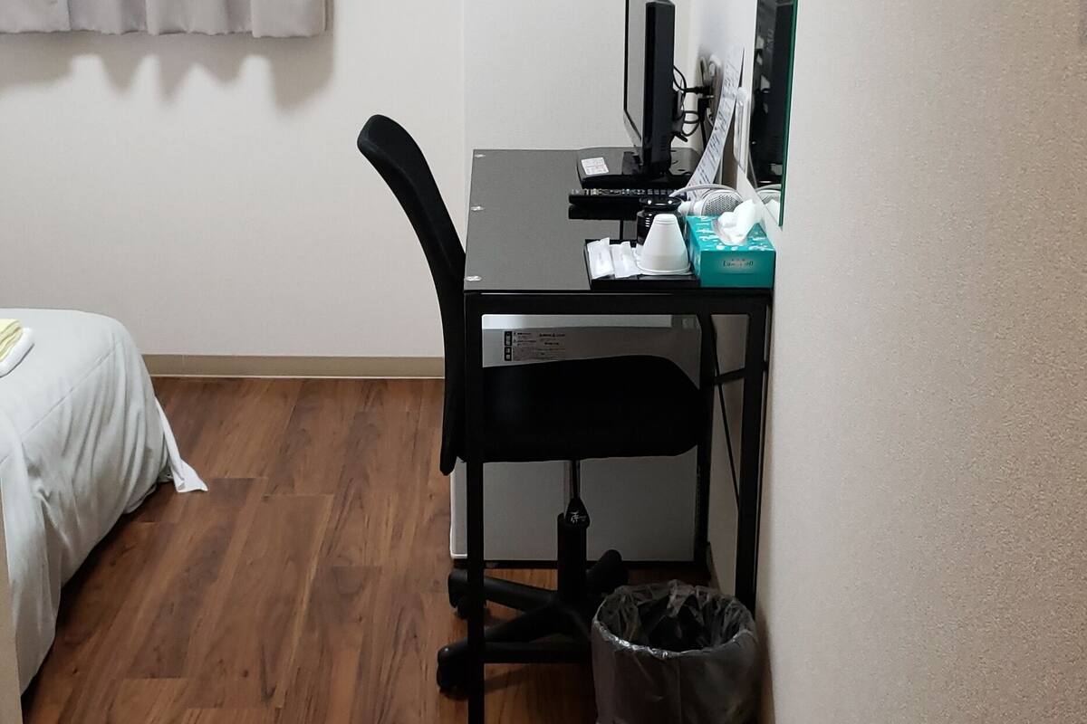 A small room with a wooden floor, a bed with white bedding, a desk with a computer, a chair, a trash bin, and a window with curtains.
