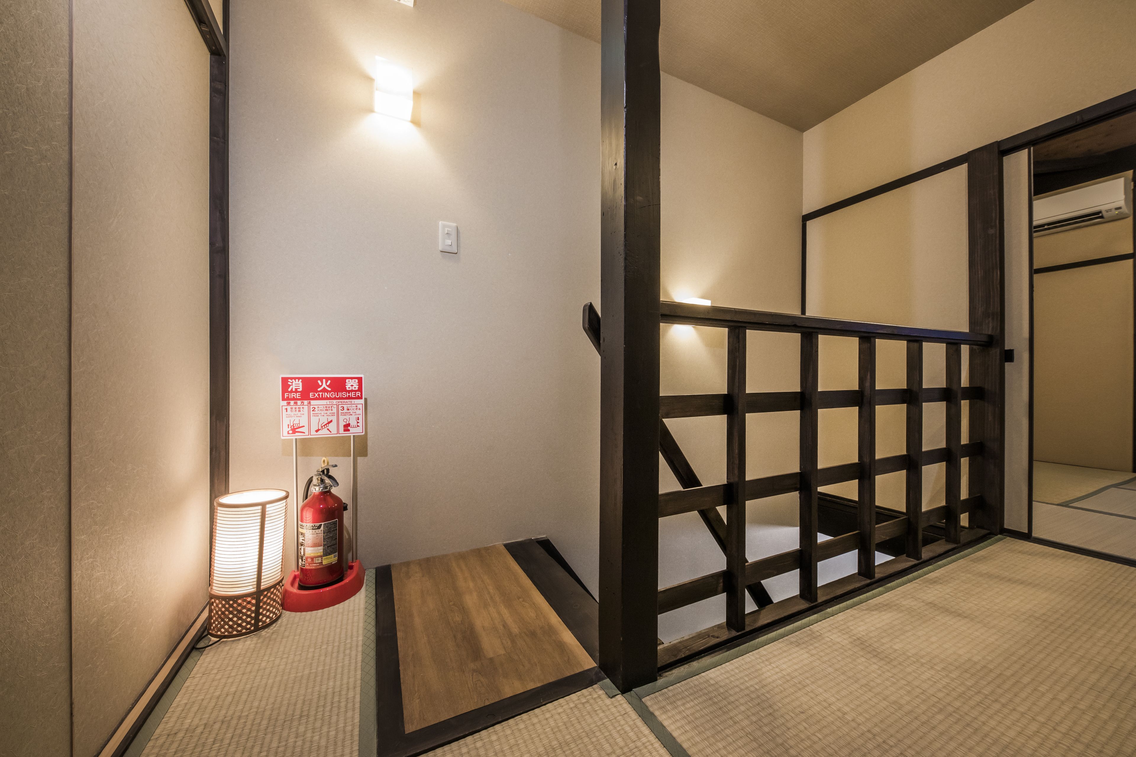 A hallway with a fire extinguisher, a lit floor lamp, and a wooden railing.