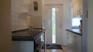 Cocina compacta con lavamanos, estufa y armarios. Se ve un ventanal con vistas a la vegetación a través de una puerta abierta.