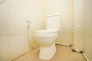 Et standard hvidt toilet i et lille, flisebelagt badeværelse med synlig rørføring og en lille smule snavs på gulvet.