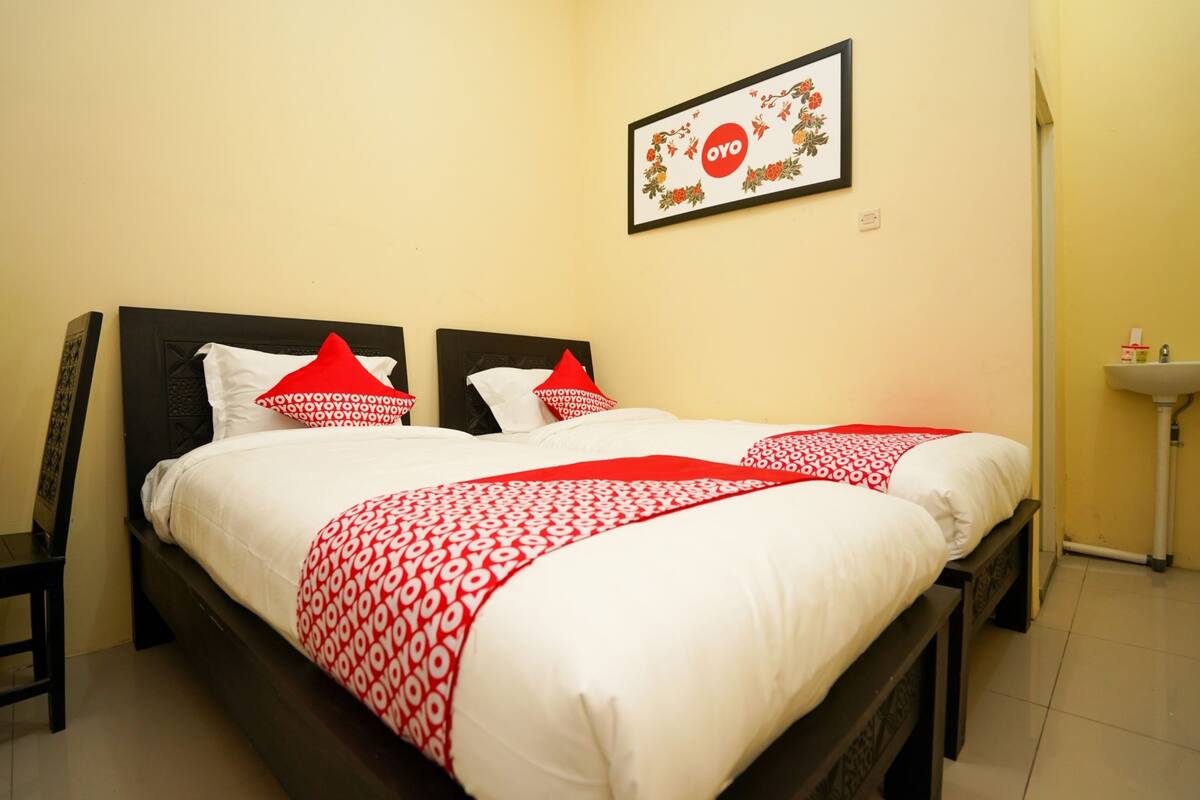 Una cama doble con ropa de cama blanca y roja, un cuadro con el texto 'OYO' y un lavamanos pequeño en la esquina.