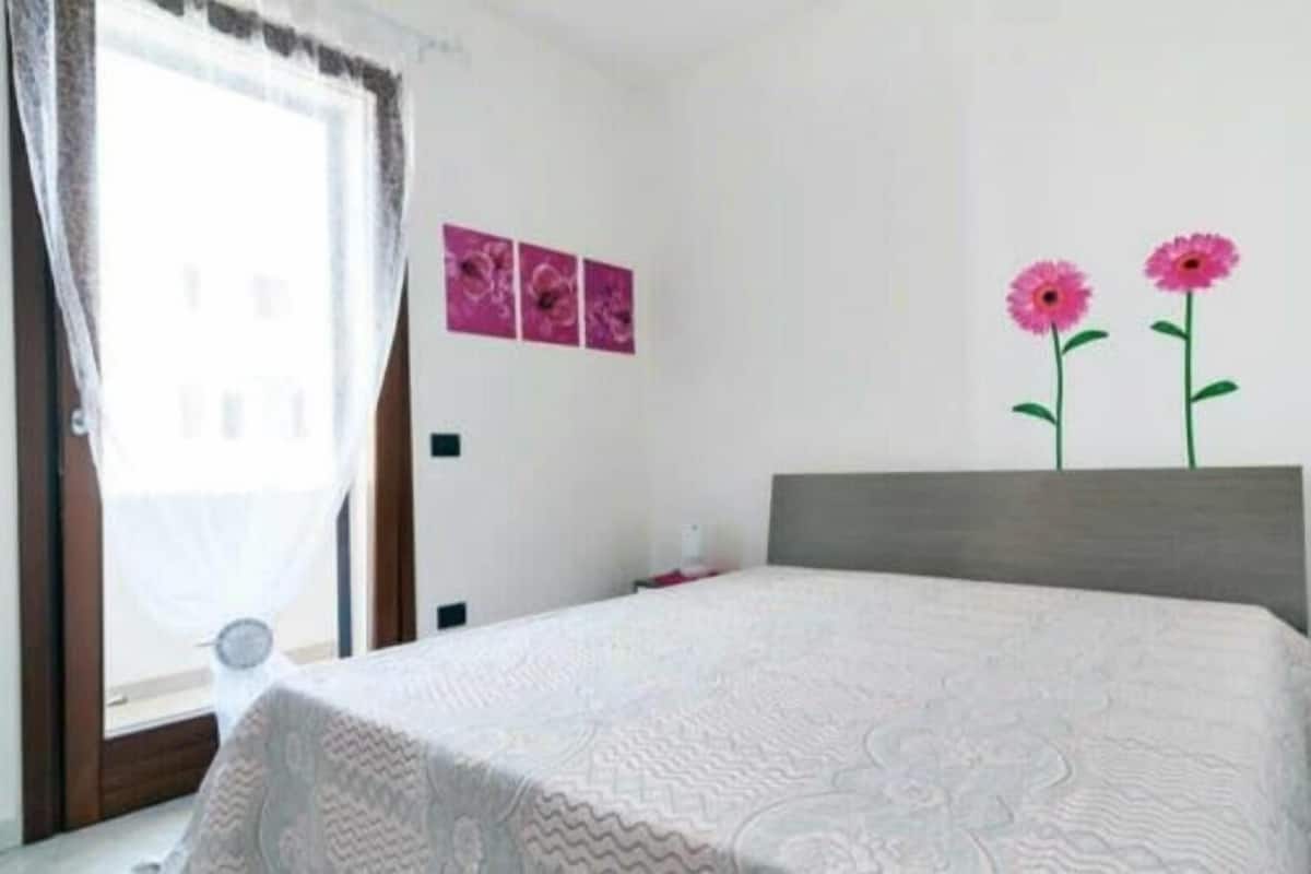 A bedroom with a bed, a window with curtains, and two pink flower decorations on the wall.