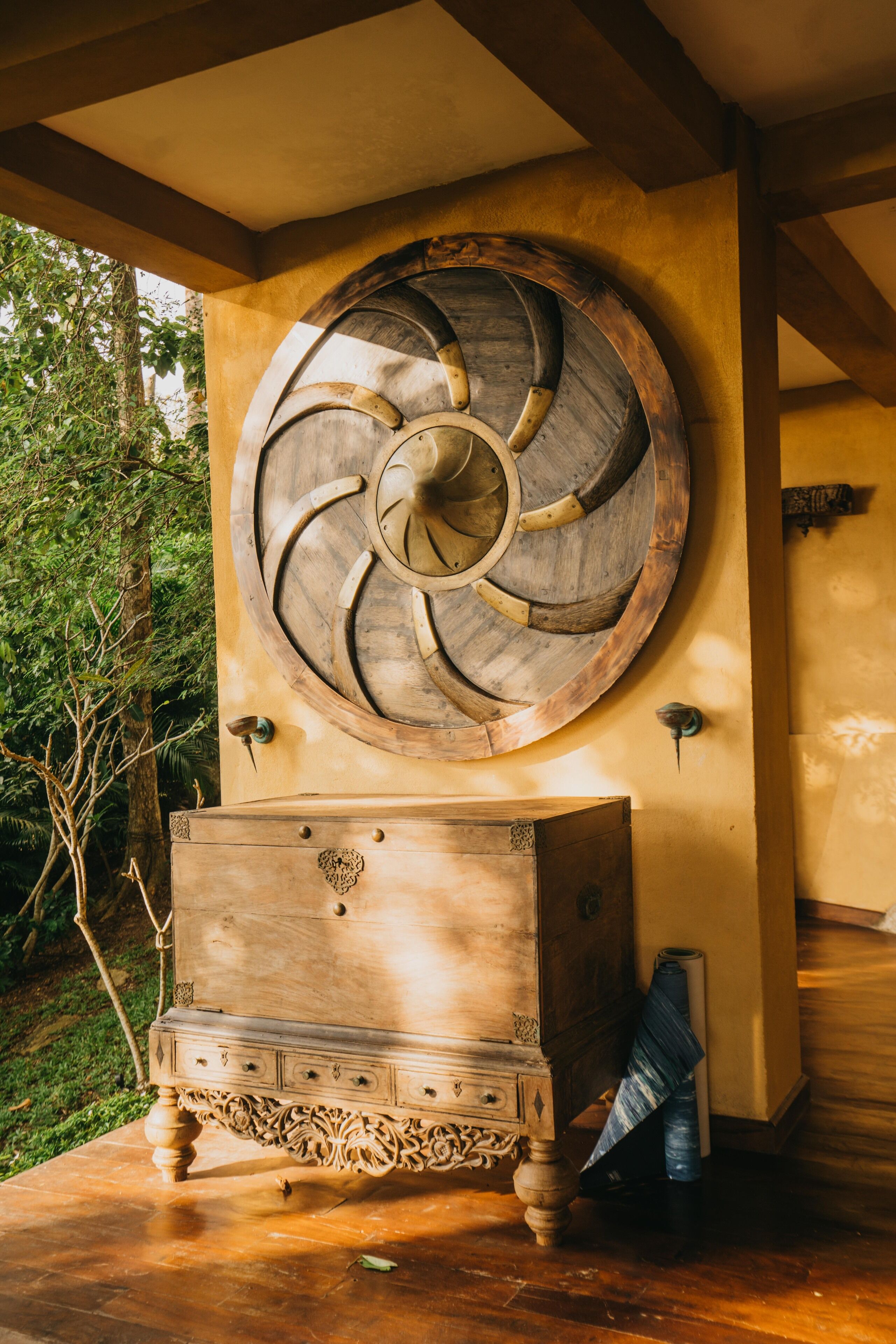 A wooden chest of drawers with intricate carvings, a large wooden wheel decoration on the wall, and a rustic wooden bench.