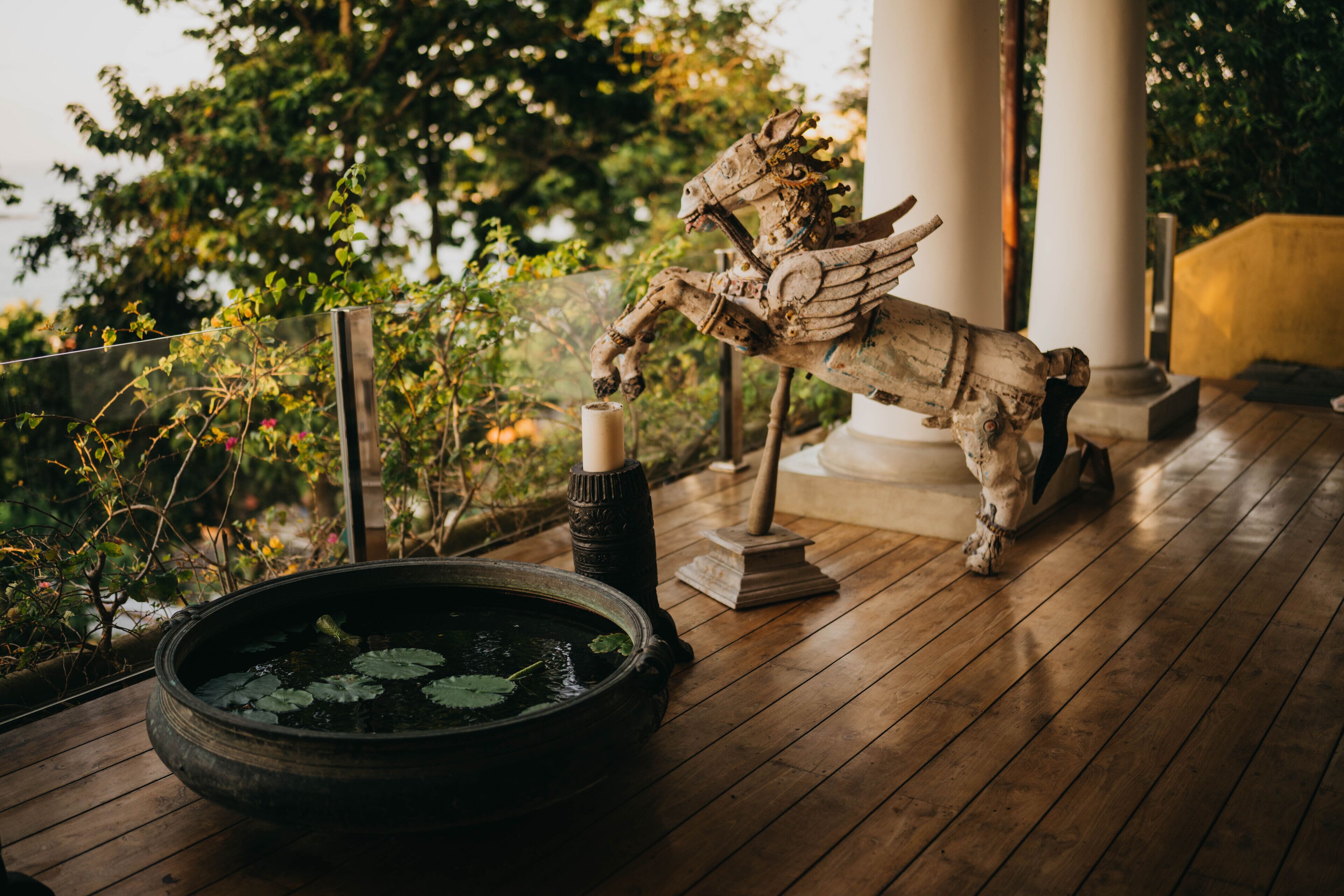 A wooden deck with a decorative horse statue, a round pond with lily pads, and a candle on a pedestal.