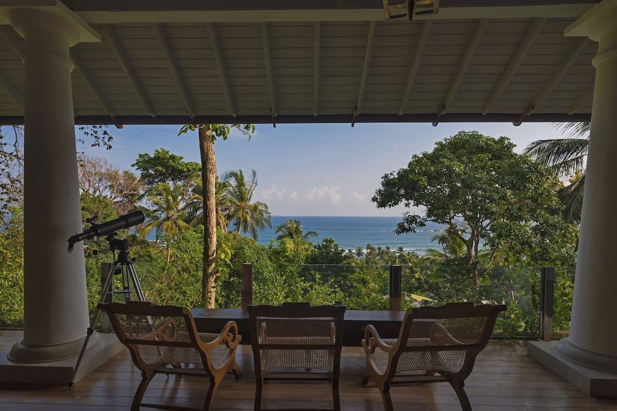 A covered outdoor seating area with a telescope, overlooking a tropical beach and ocean.