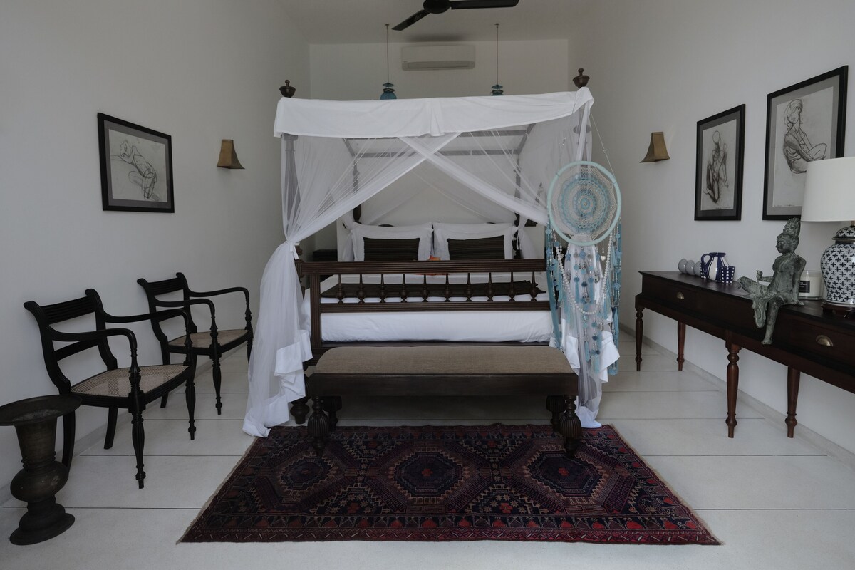 A bedroom with a four-poster bed, a canopy, a patterned rug, a nightstand, a lamp, and framed artwork on the walls.