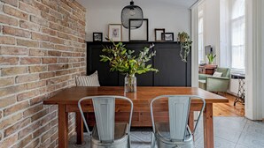 Un espace repas avec un mur en briques, une table en bois, des chaises en métal, un luminaire suspendu et un fond noir agrémenté de tableaux encadrés et de plantes.