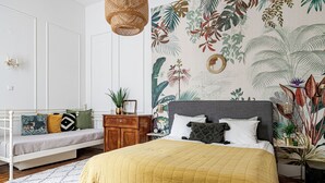 Une chambre à coucher comprenant un lit, une table de chevet, une commode, une chaise et un mur orné d’une fresque représentant des plantes tropicales.