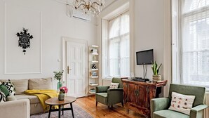 Un salon avec un canapé, des fauteuils, une table basse et un téléviseur. On y trouve également une bibliothèque et une plante.