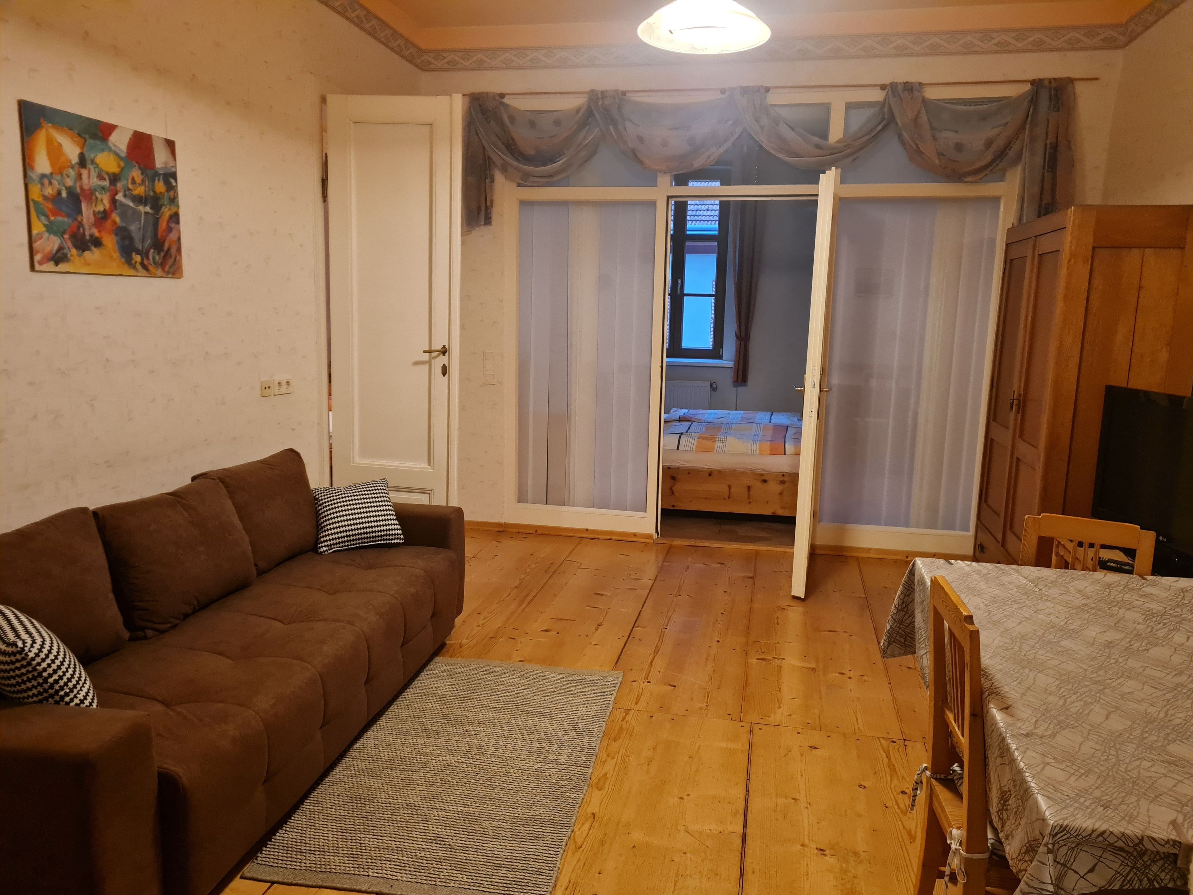 Huoneessa on ruskea sohva, pieni pöytä, puinen päätyosaan varustettu sänky, seinällä oleva taulu ja ovi, joka johtaa toiseen huoneeseen.