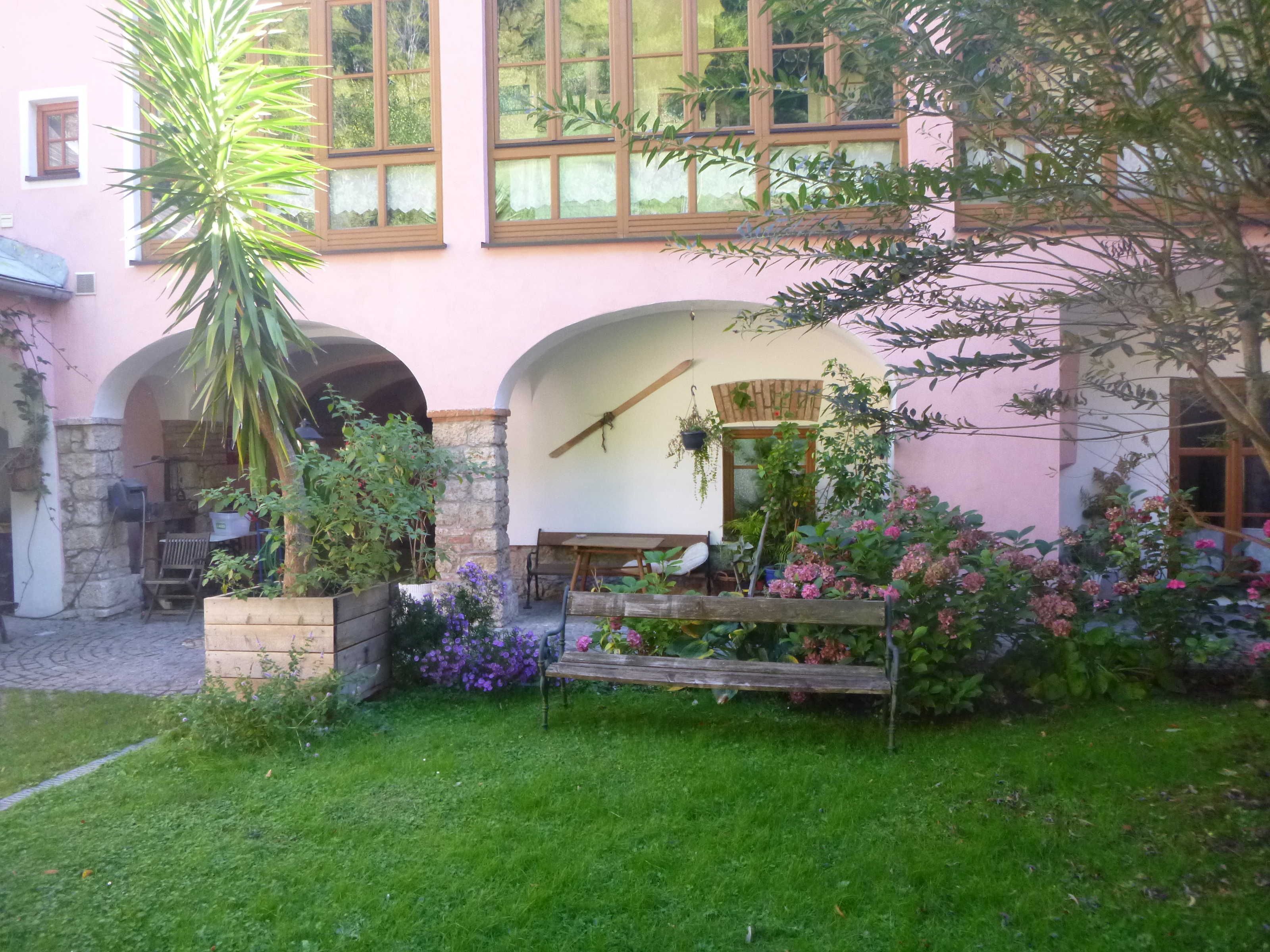 Un bâtiment rose avec un jardin à l’avant, comprenant un banc et des plantes en pot.