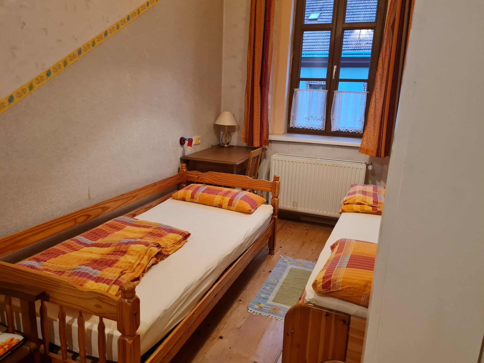 Ein kleiner, gemütlicher Raum mit zwei hölzernen Betten, einem Nachttisch, einer Lampe und einem Fenster mit Vorhängen.