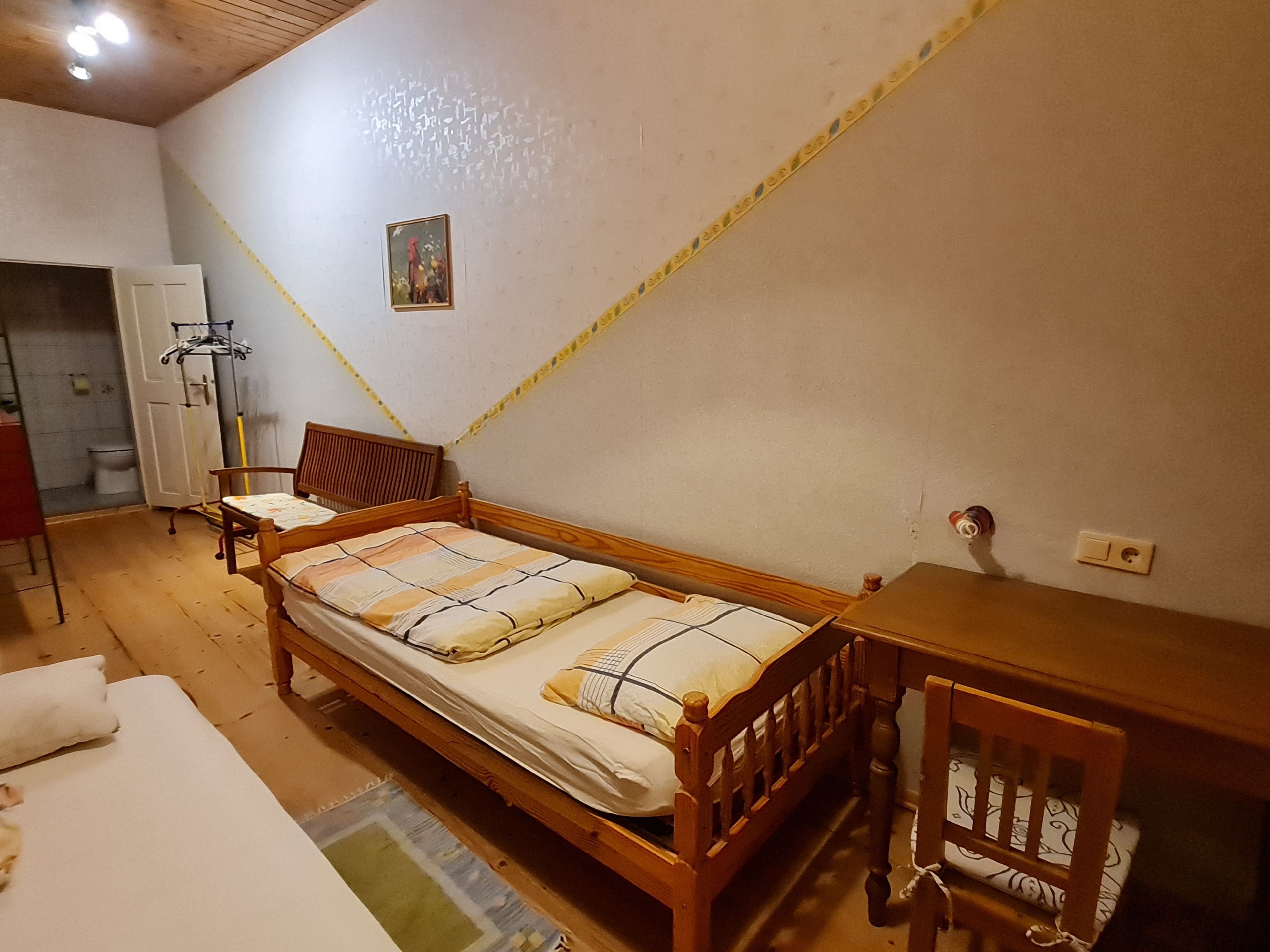 A single bed with a wooden frame, a small bedside table, a chair, and a wardrobe in a room with wooden flooring and a sloped ceiling.