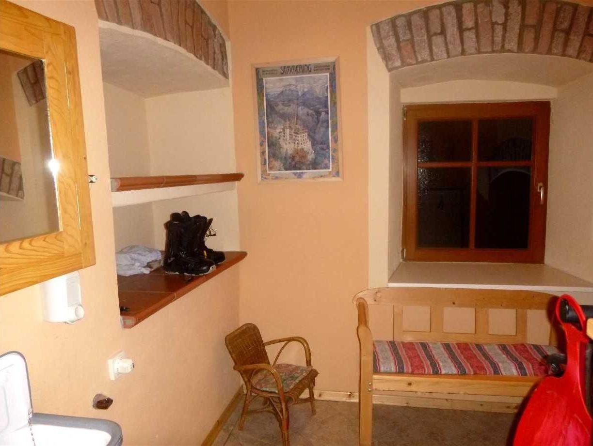 Een kleine kamer met een houten bankje, een stoel, een plank met schoenen, een raam en een poster aan de muur.