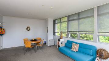 A modern living room with a blue sofa, a dining table with chairs, and large windows with blinds.