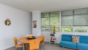 Una sala de estar moderna con una mesa redonda, sillas anaranjadas, un sofá azul y ventanas amplias con persianas.