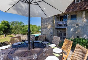 A round wooden table with chairs, a large umbrella, and a stone building with a balcony in the background. - Stunning, family-friendly farmhouse; large garden, safe private pool (Rueyres)
