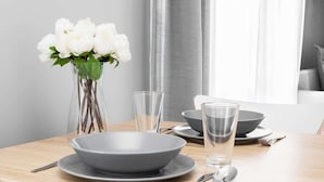 Uma mesa de jantar com uma tigela cinza, dois copos transparentes e um vaso com flores brancas.