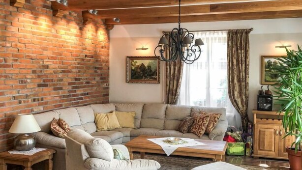A cozy living room with exposed wooden beams, a brick wall, a sectional sofa, a chandelier, and a large window with curtains. - Zielona Dolina Młyn by Interhome (Kwilcz)