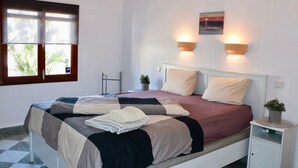 Quarto com cama, duas luminárias na parede, janela com persianas, uma planta pequena em uma mesa de cabeceira e um quadro na parede.
