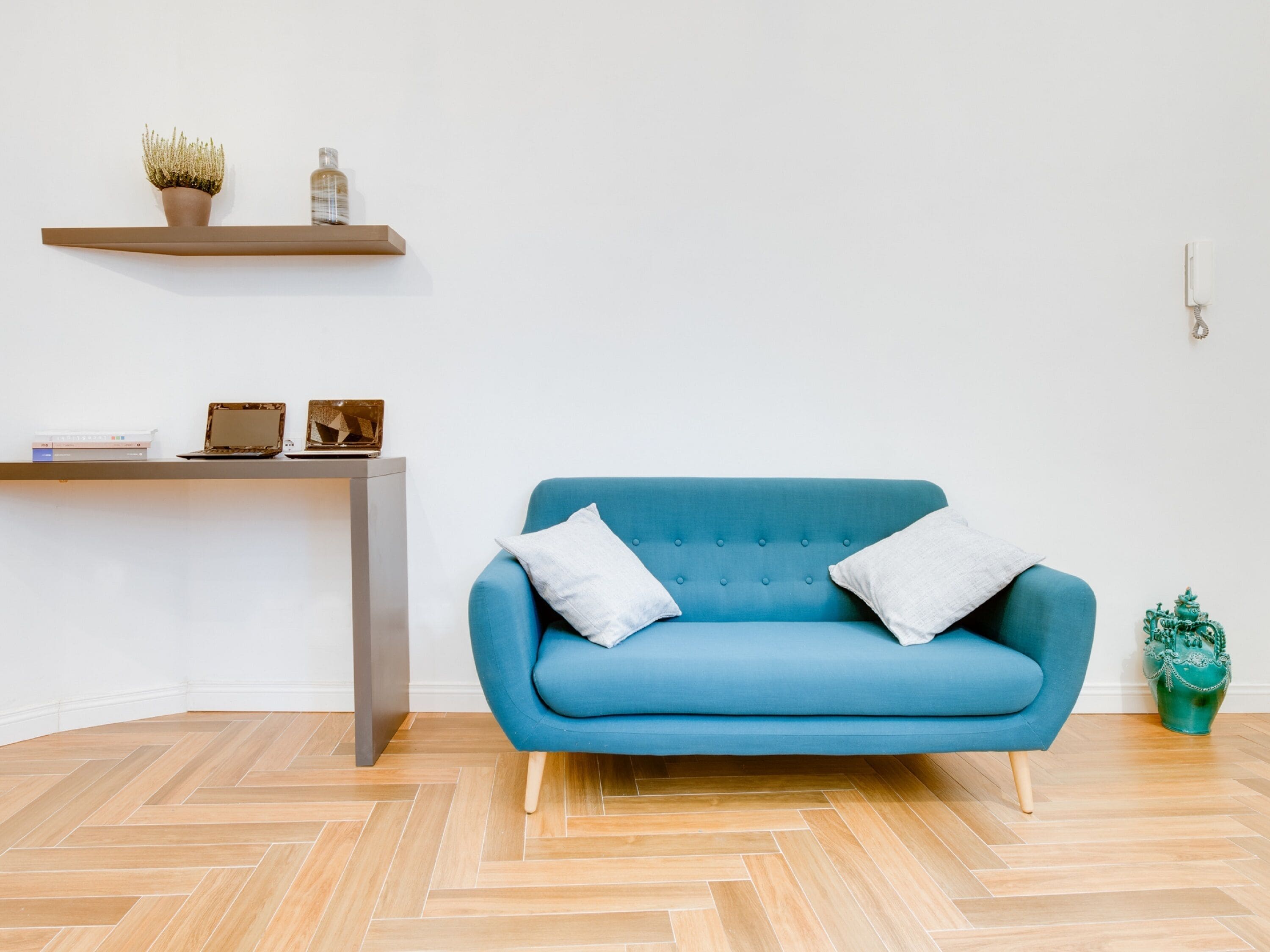 A blue sofa with two pillows, a wooden herringbone floor, a small desk with a laptop, a vase, and a telephone on the wall.