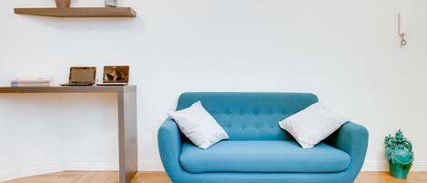 A blue sofa with two pillows, a wooden herringbone floor, a small desk with a laptop, a vase, and a telephone on the wall.