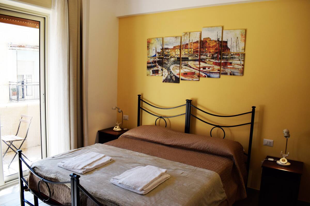 Quarto de hotel com uma cama, mesa de cabeceira, abajur e janela com vista para uma varanda.