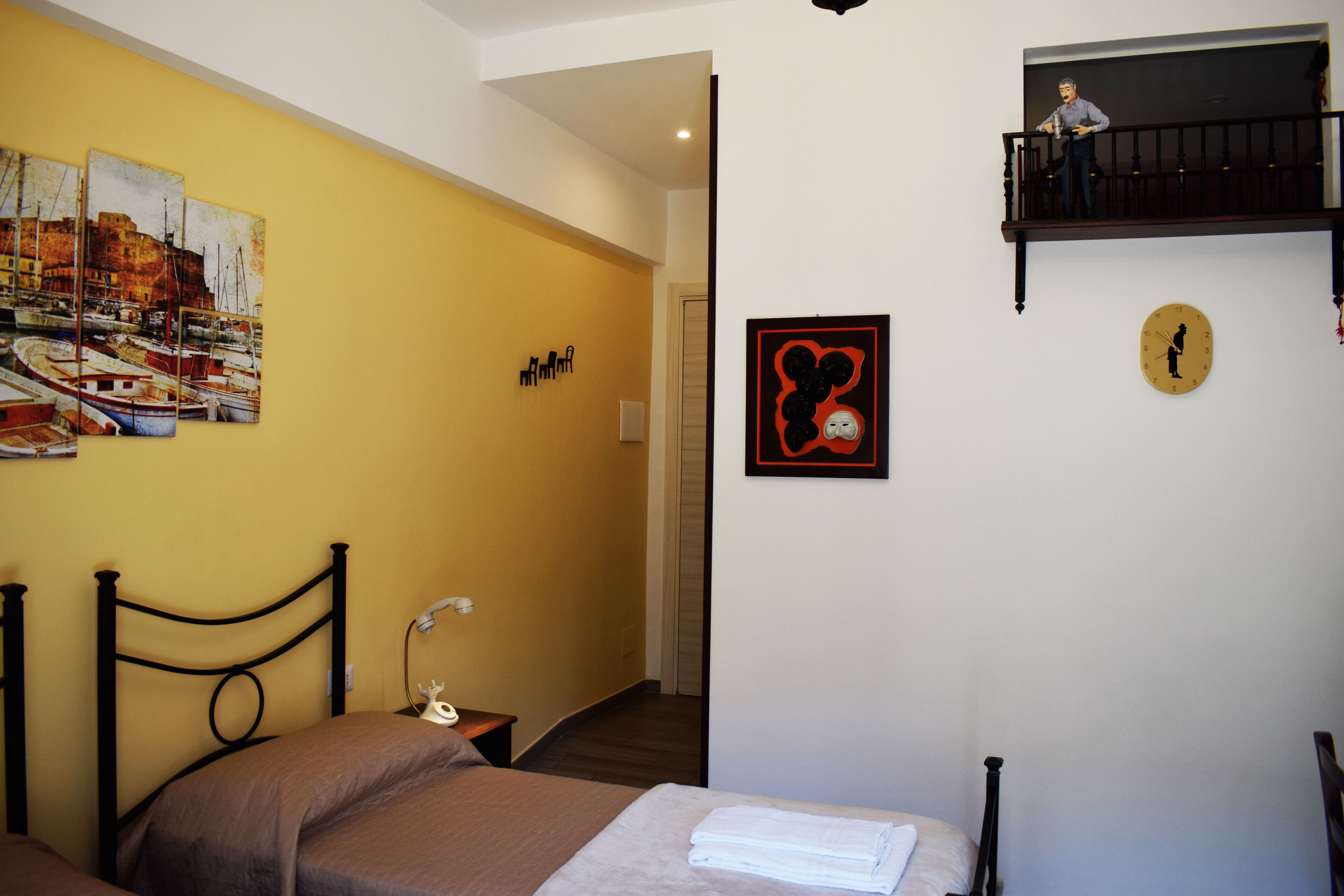 Une chambre d’hôtel avec un lit, une table de chevet, une lampe, un tableau représentant des bateaux, une horloge et un petit balcon.