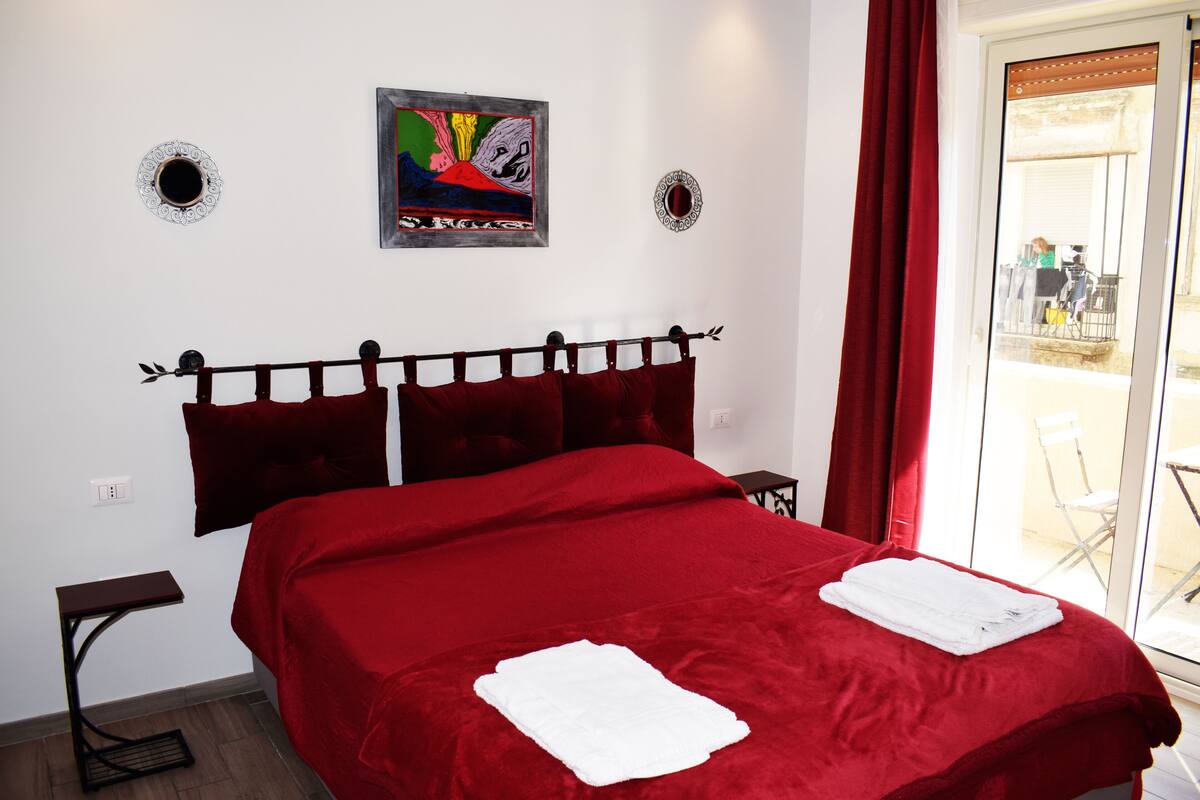 Quarto de hotel com uma cama grande, roupa de cama vermelha, duas almofadas, uma cabeceira, uma mesinha de cabeceira e uma janela com cortinas vermelhas.