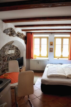 Quarto com lareira de pedra, uma cama, escrivaninha com cadeira e duas janelas com cortinas.