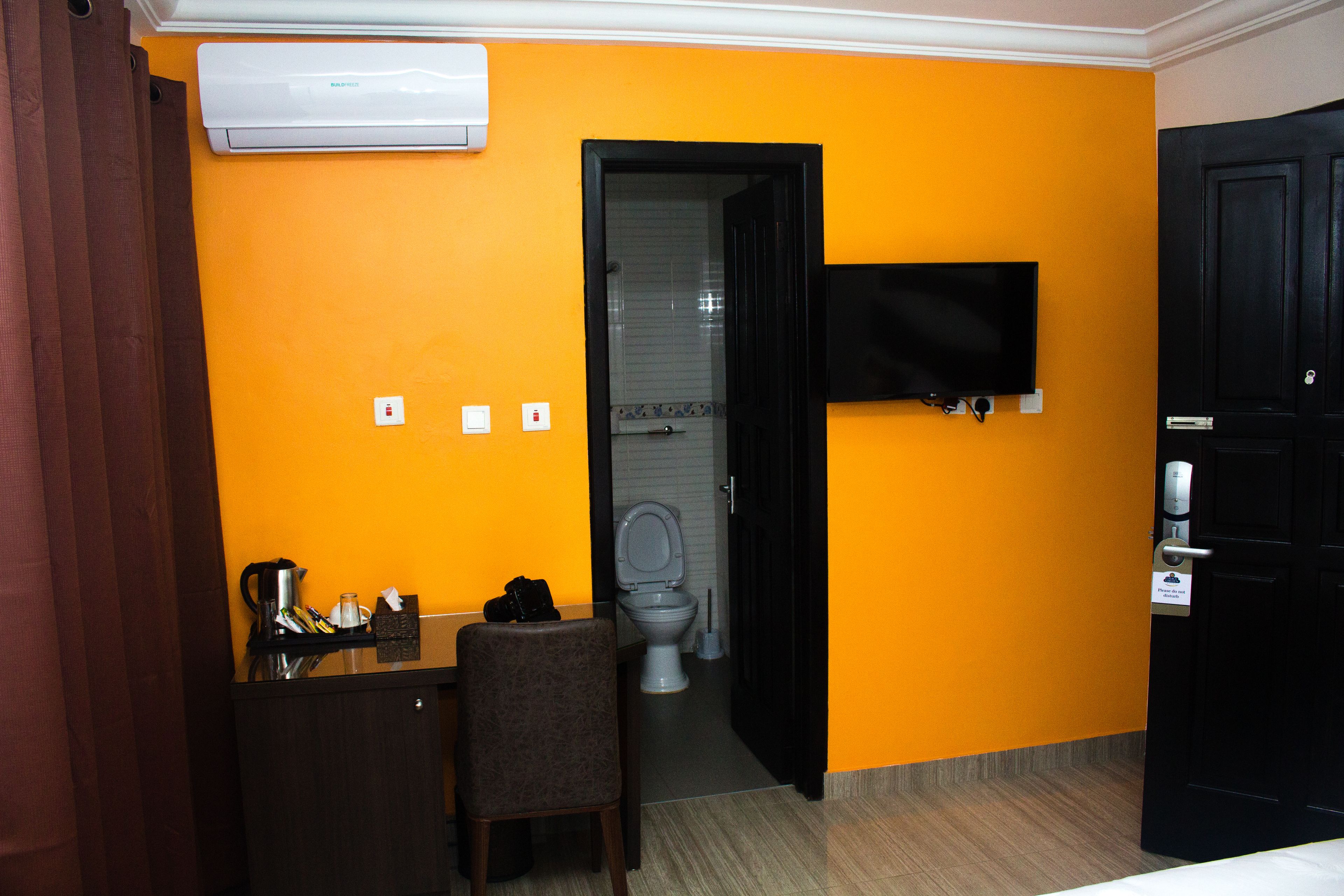 Habitación de hotel con una pared naranja, una puerta negra, un televisor montado en la pared, un escritorio con una silla y un inodoro visible a través de una puerta abierta.
