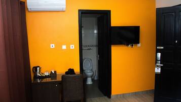 Habitación de hotel con una pared naranja, una puerta negra, un televisor montado en la pared, un escritorio con una silla y un inodoro visible a través de una puerta abierta.