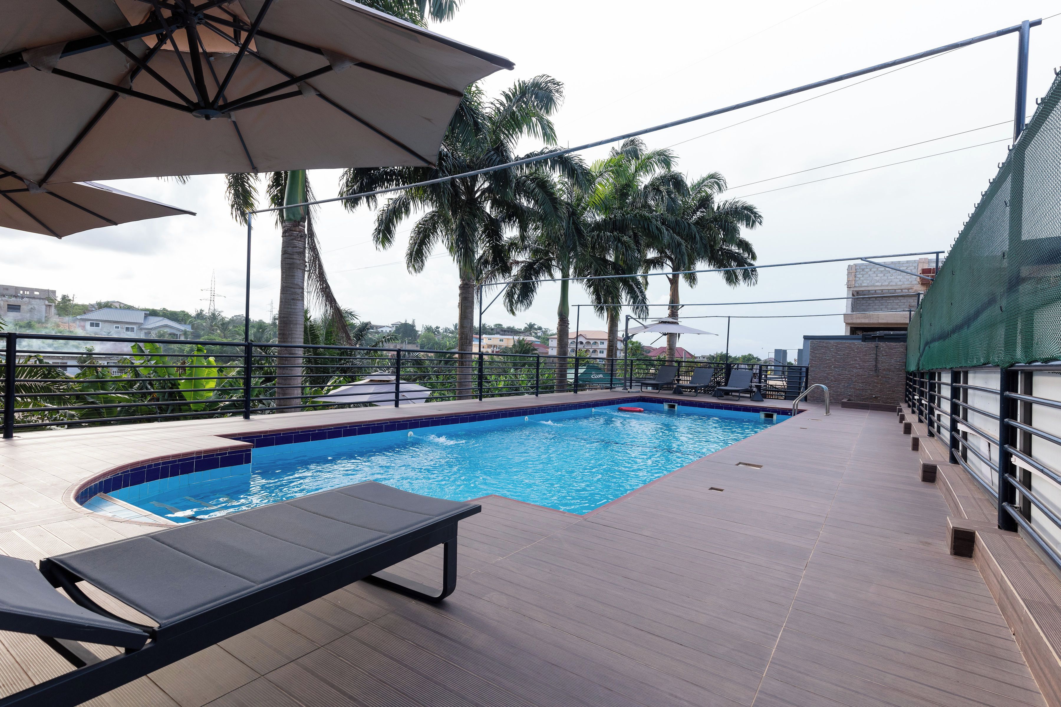 Una piscina con sillones de descanso, rodeada de palmeras y vista a edificios al fondo.