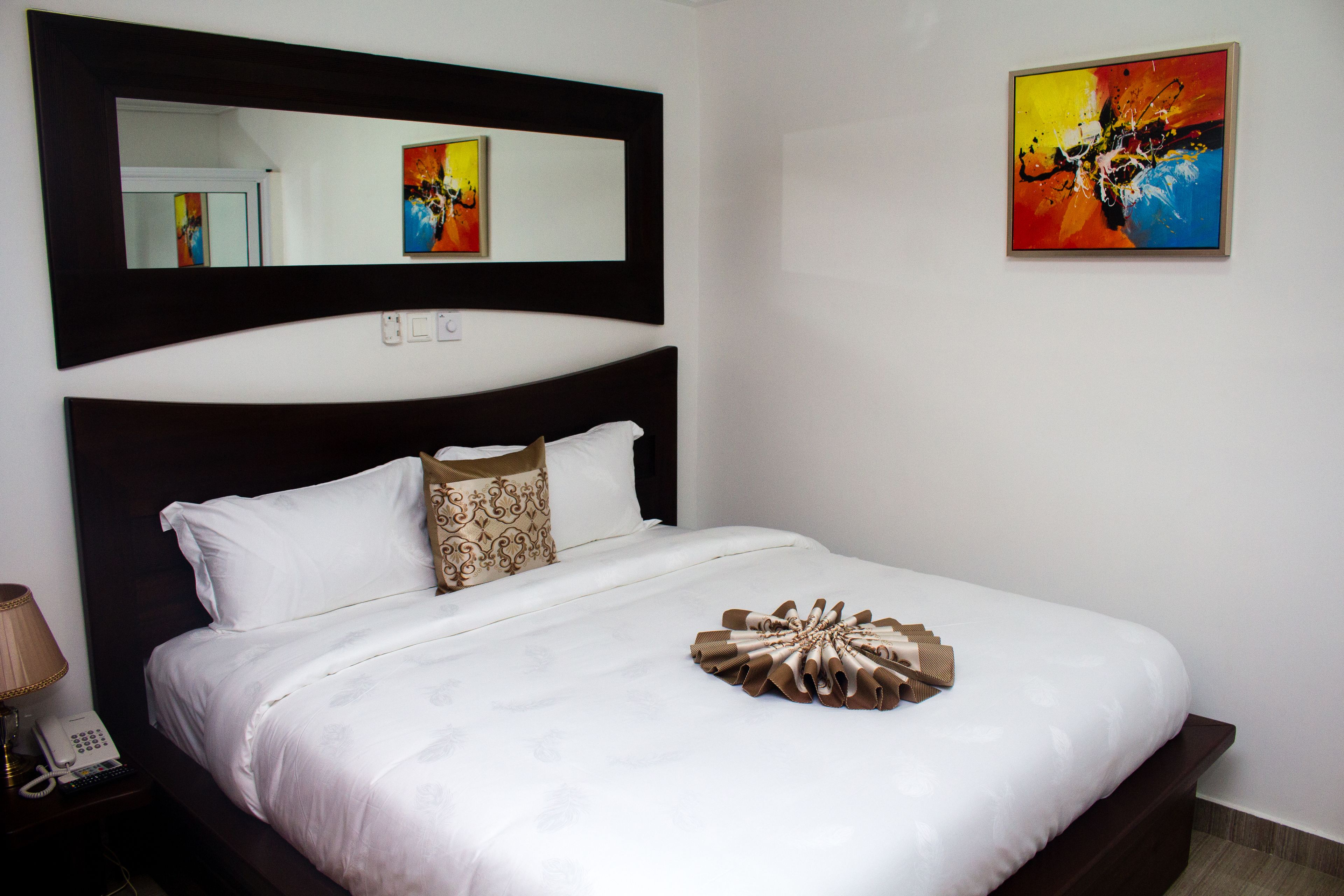 A neatly made bed with white linens and decorative pillows, a bedside telephone, and a framed abstract painting on the wall.