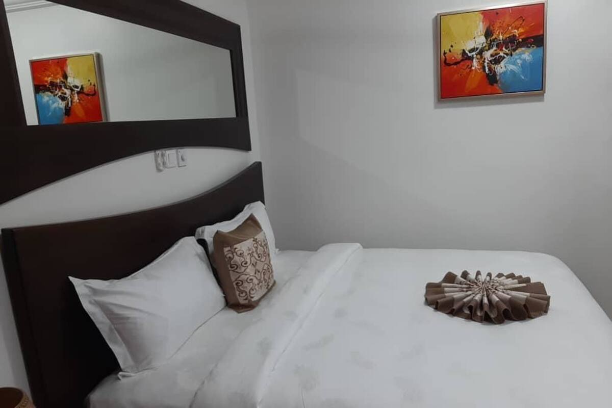 Una cama bien hecha con ropa blanca y un cojín decorativo, un espejo sobre la cama y dos cuadros en la pared.