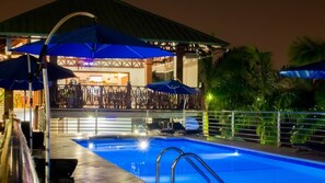 Una piscina con zona cubierta y sombrillas, rodeada de palmeras y un edificio con bar.
