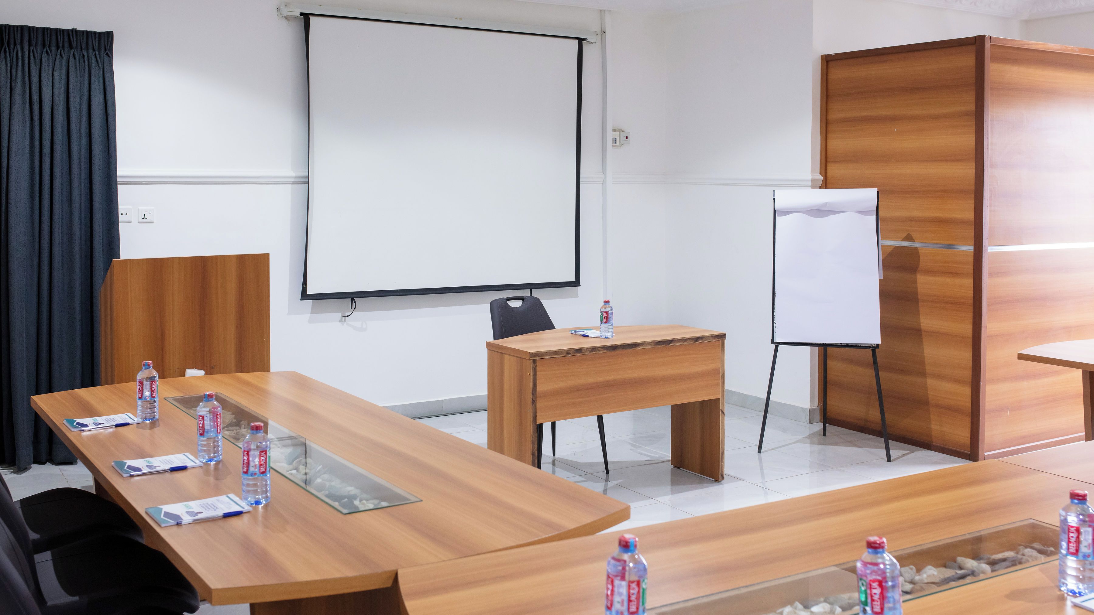 Une salle de conférence équipée d’une longue table en bois, d’un tableau blanc, d’un écran de projection et d’un réfrigérateur blanc.