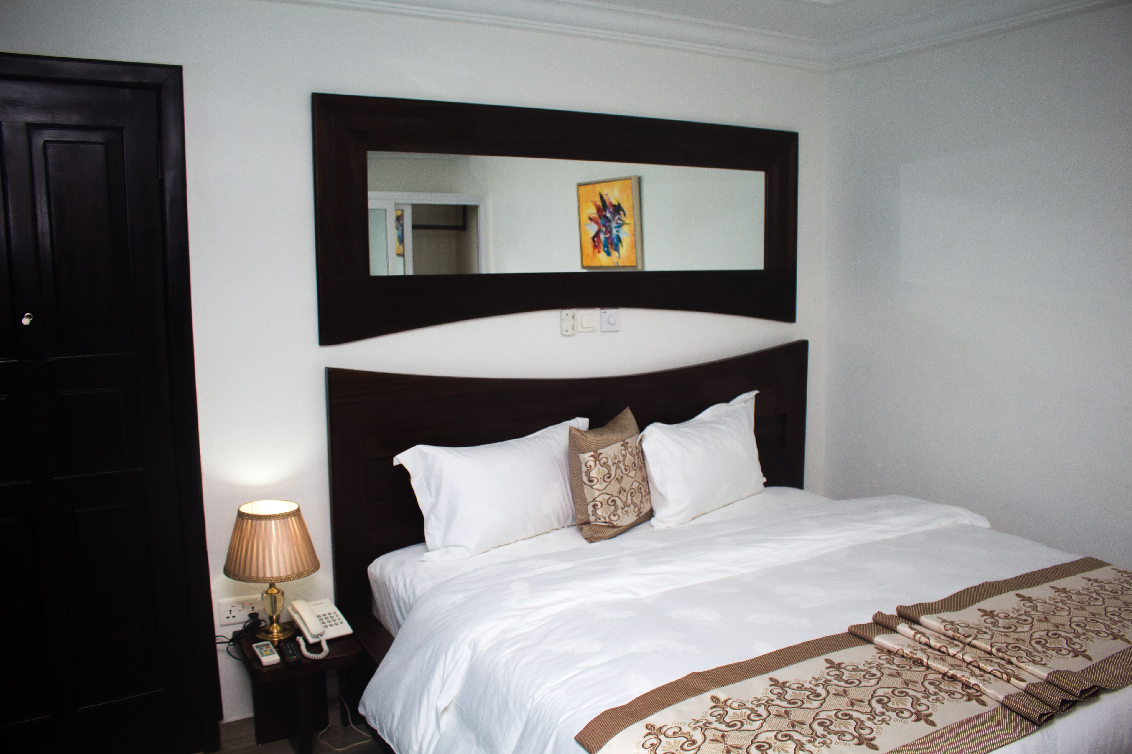 Habitación de hotel con cama, lámpara de noche, teléfono, espejo y un cuadro en la pared.