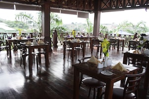 A restaurant with wooden tables and chairs, arranged in a spacious outdoor area with a view of palm trees and buildings.
