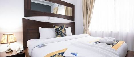 A hotel room with a large bed, a nightstand with a lamp, a mirror, and a window with curtains.