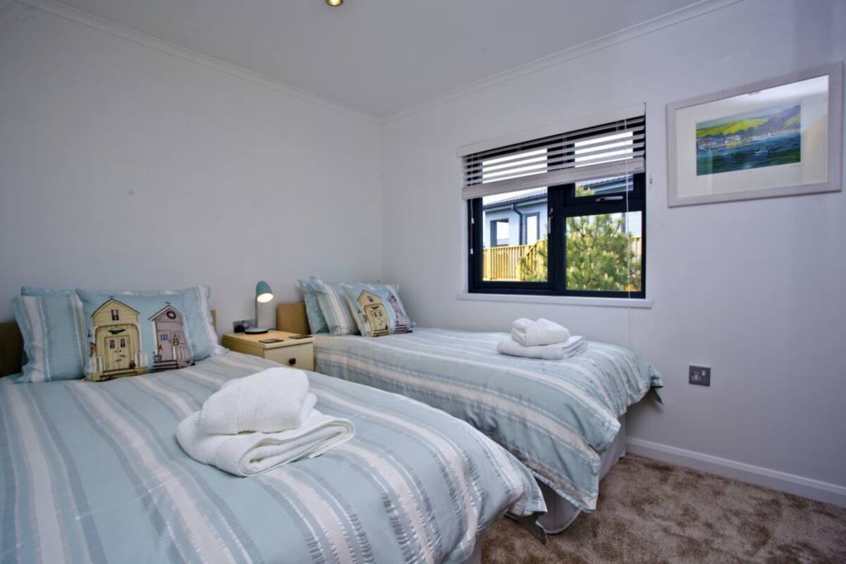 A bedroom with two beds, a nightstand, a lamp, a window with blinds, and a framed picture on the wall.